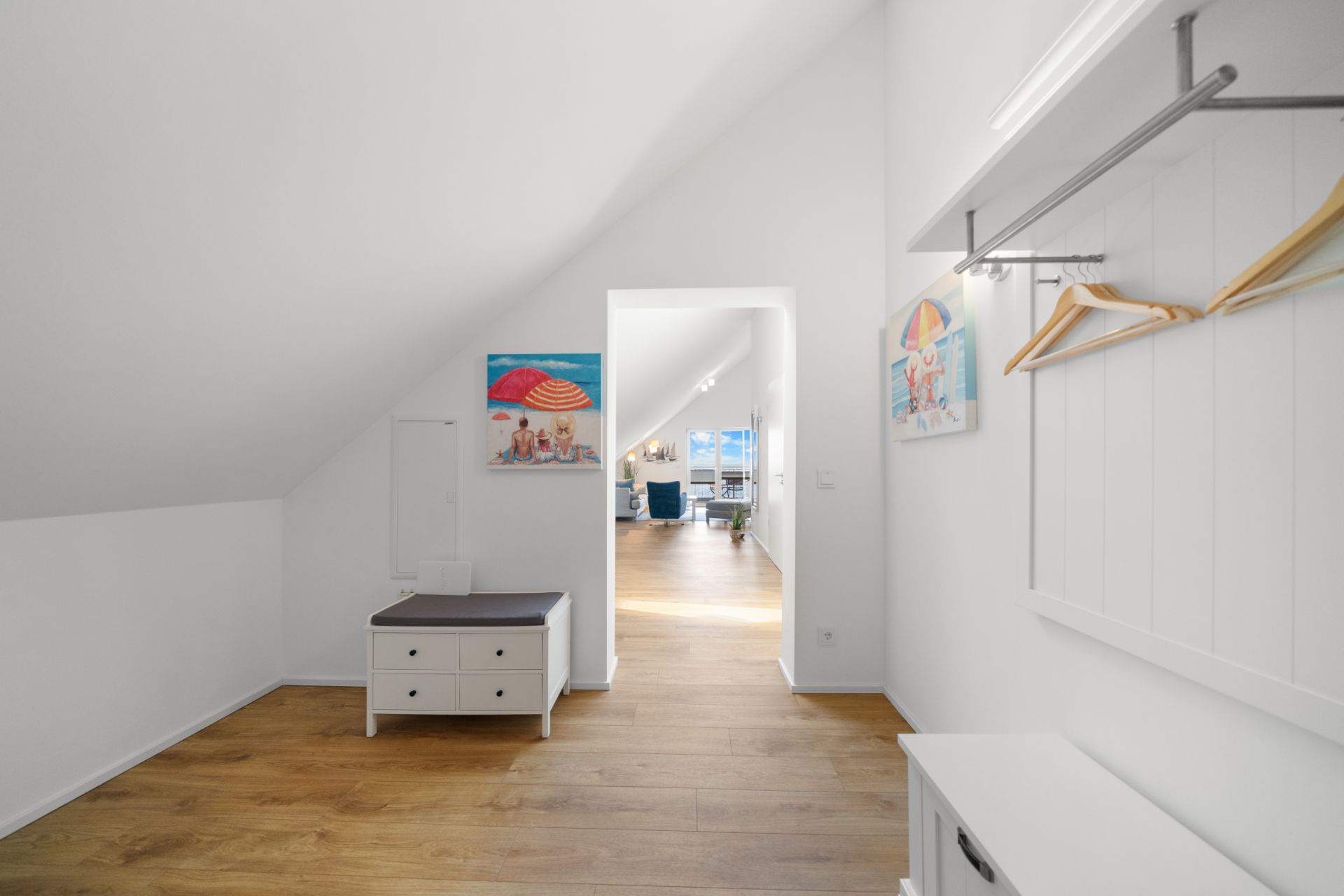 Heller Flur mit Holzboden, Sitzbank, Kleiderhaken und bunten Bildern, Blick ins Wohnzimmer.