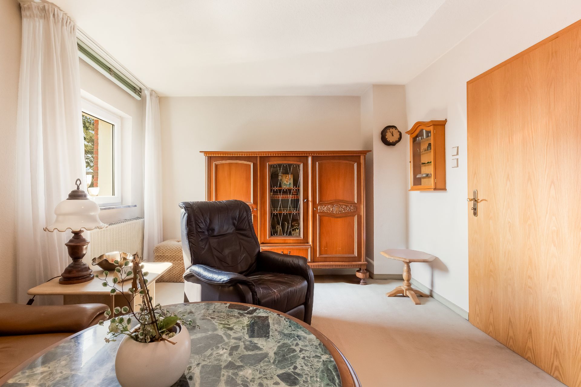Helles Wohnzimmer mit Ledersessel, Holzmöbeln, Marmortisch und großem Fenster.