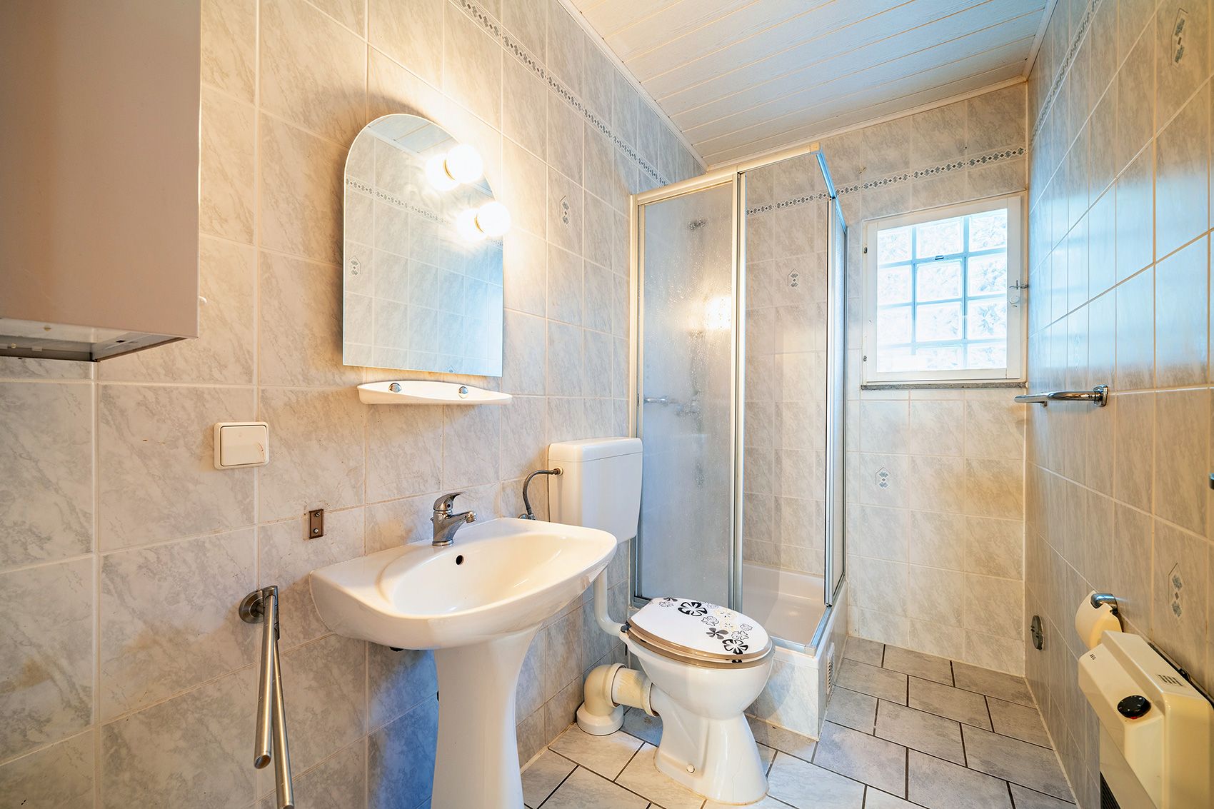 Helles Badezimmer mit Waschbecken, Spiegel, Toilette und Dusche, gefliest und mit Fenster.