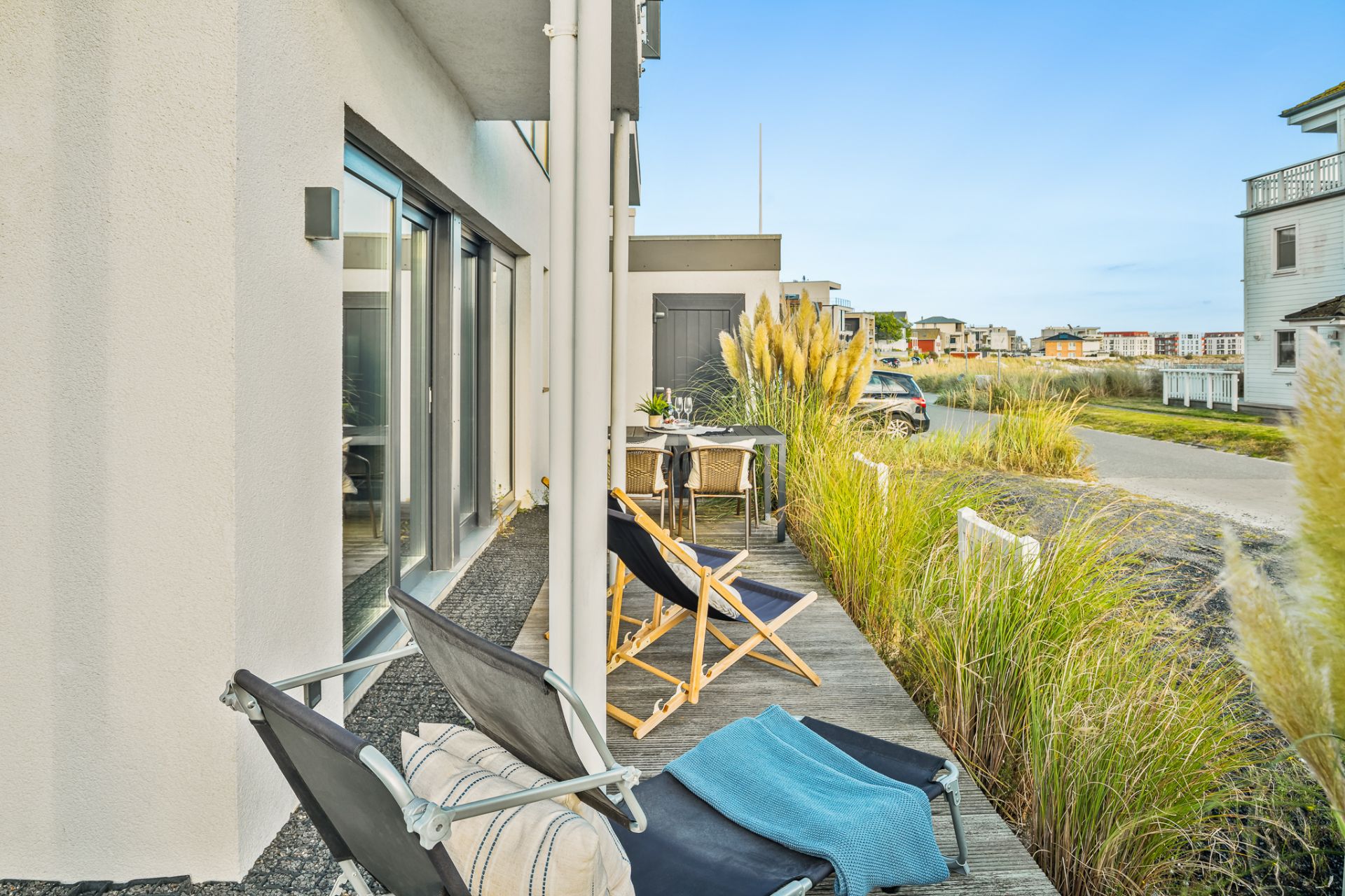 Terrasse mit Liegestühlen, Tisch und Stühlen neben einem Wohnhaus, umgeben von Ziergras.
