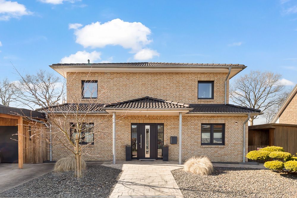 Modernes zweistöckiges Haus mit hellem Klinker und schwarzem Dach, Vorgarten mit Steinbeeten und kleinen Büschen.
