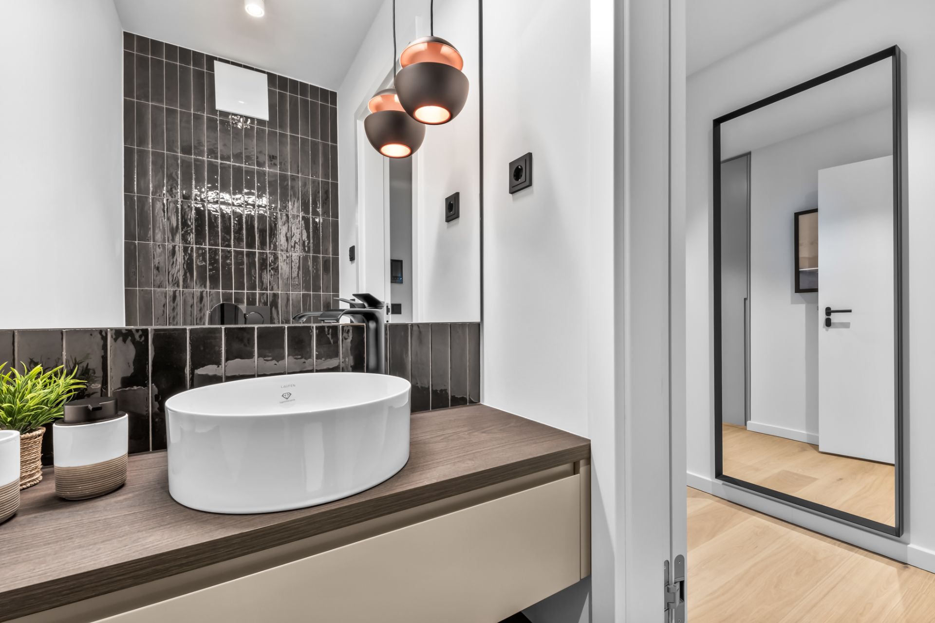 Modernes Badezimmer mit rundem Waschbecken, dunklem Fliesenspiegel und zwei hängenden Lampen.