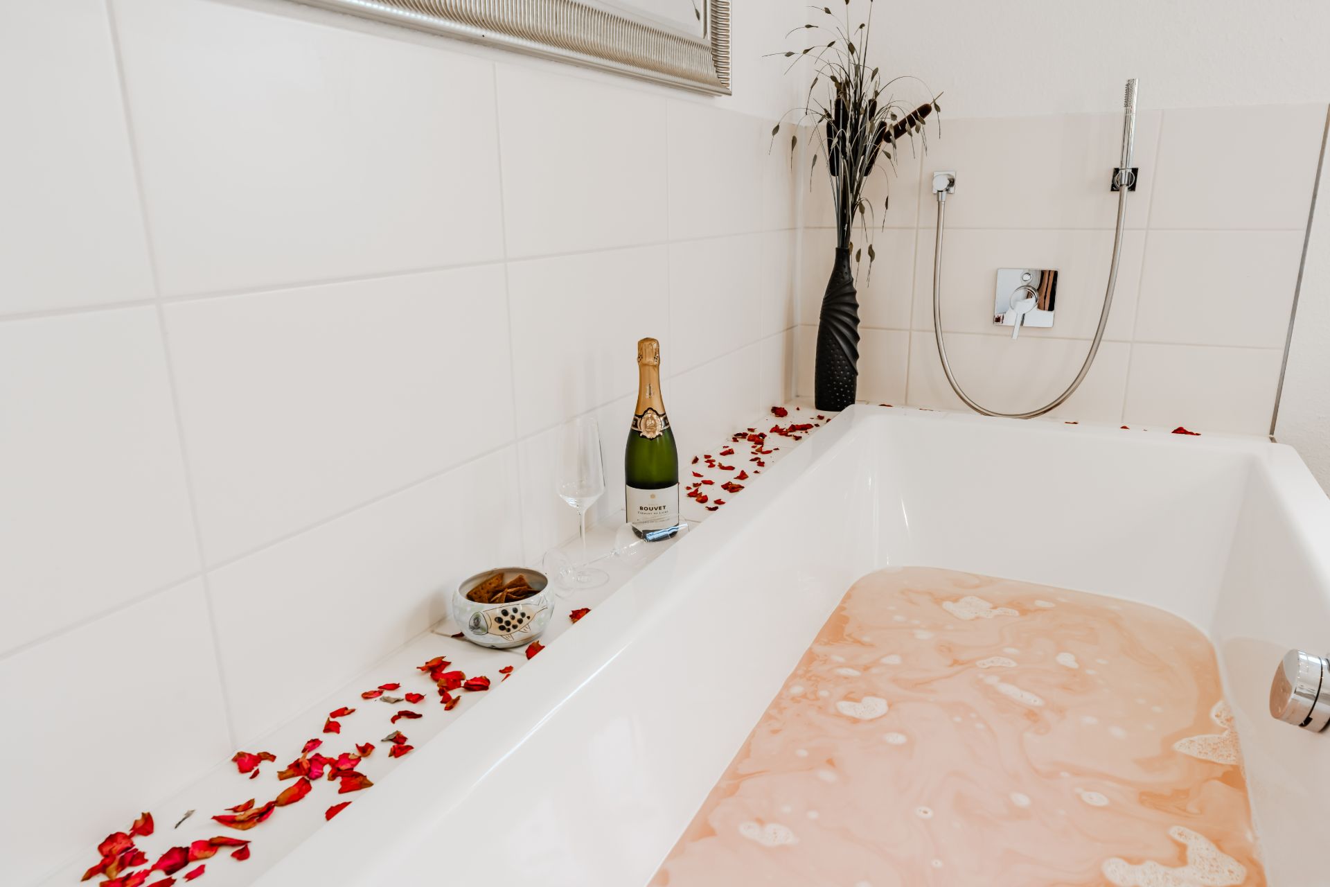 Weiße Badewanne mit Wasser, Rosenblättern, Sektflasche, Glas und Vase mit Zweigen auf dem Wannenrand.