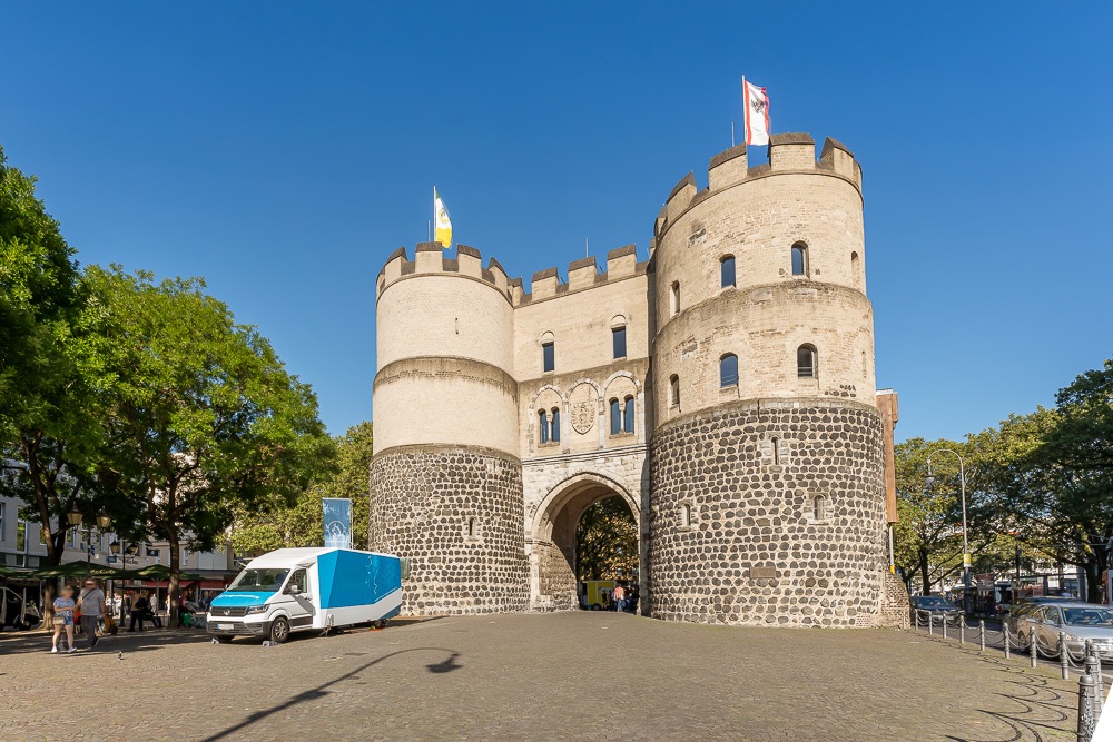 Altes Steintor mit zwei runden Türmen, blau-weißer Lieferwagen davor, blauer Himmel.