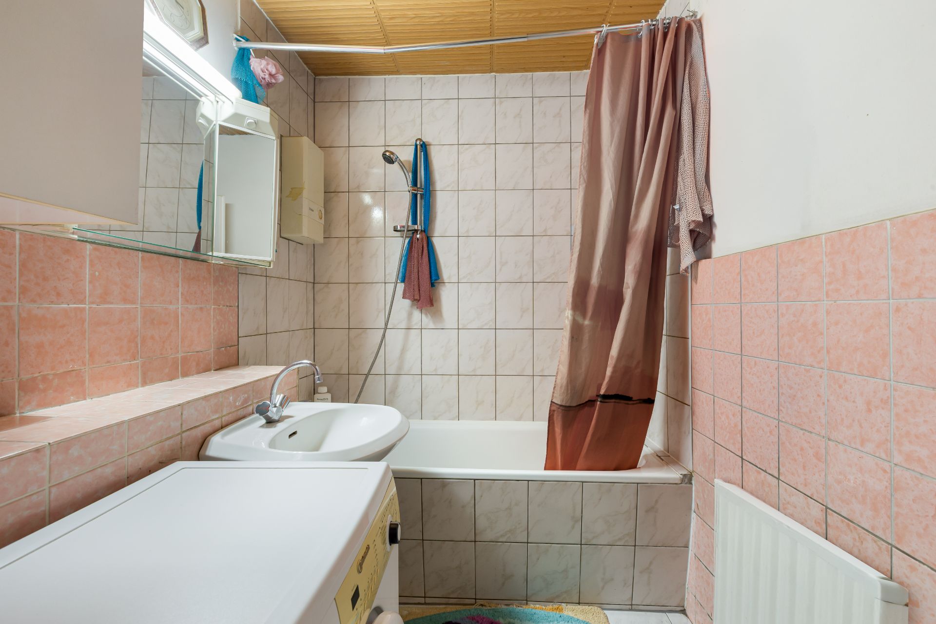 Badezimmer mit Badewanne, Waschbecken, Waschmaschine und Duschvorhang, gefliest mit rosa Kacheln.