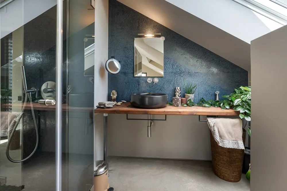 Modernes Badezimmer mit schräger Decke, Holzwaschtisch, schwarzem Waschbecken und Pflanzen.