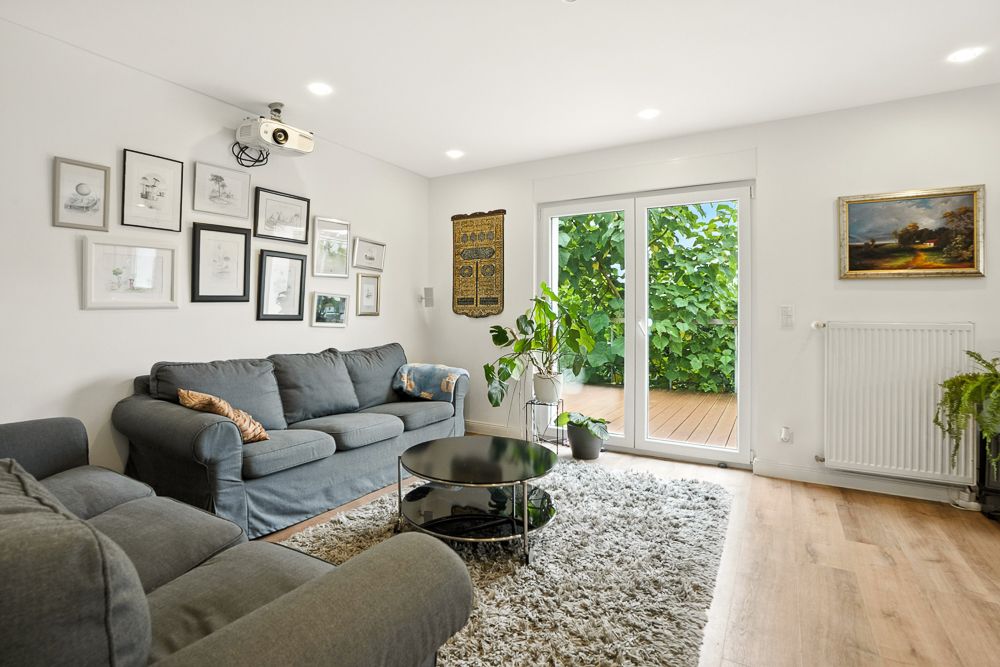 Helles Wohnzimmer mit zwei grauen Sofas, Teppich, Glastisch, Pflanzen und Wandbildern.