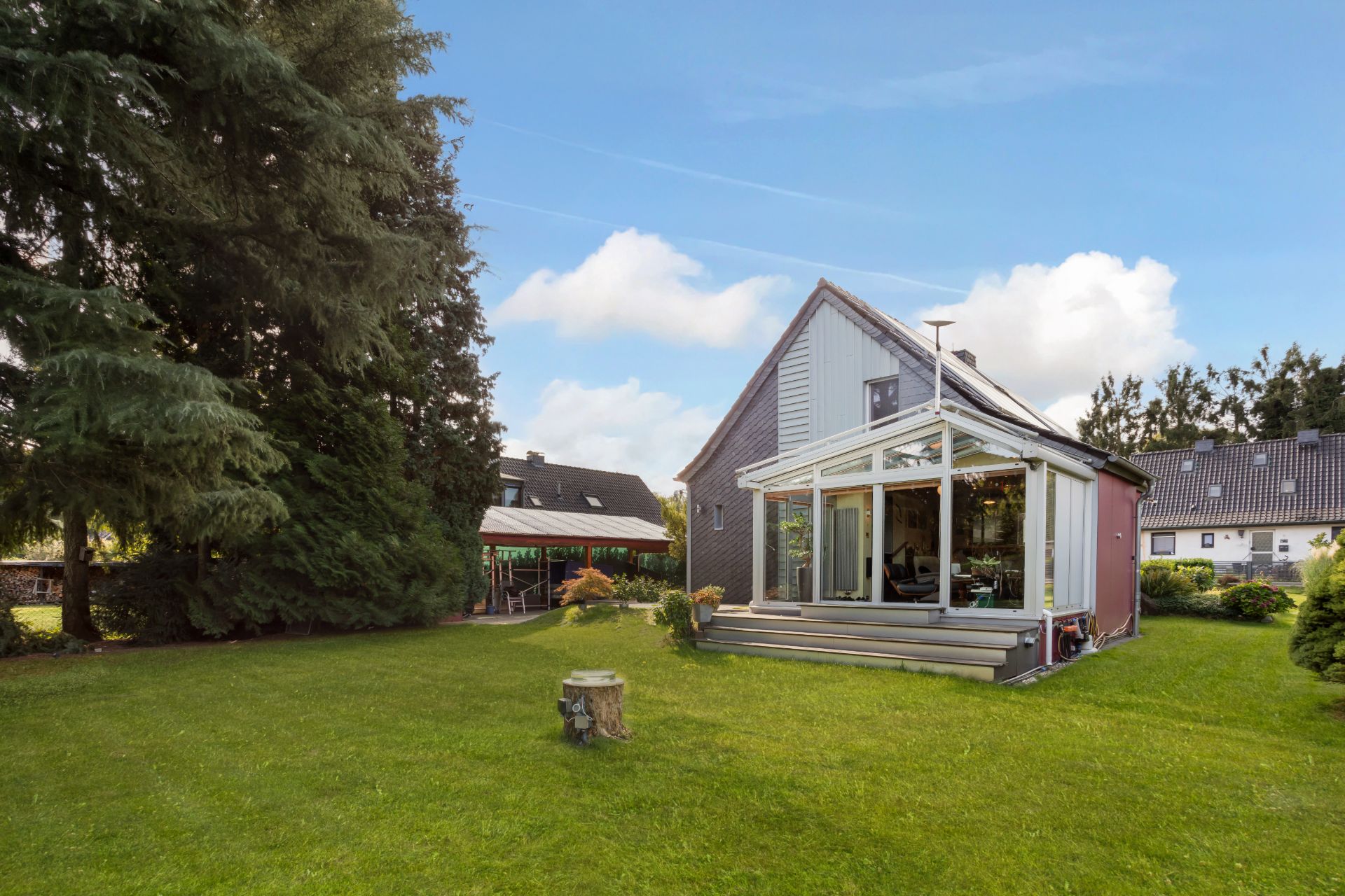 Haus mit Wintergarten und großer Wiese, umgeben von Bäumen und Nachbarhäusern.