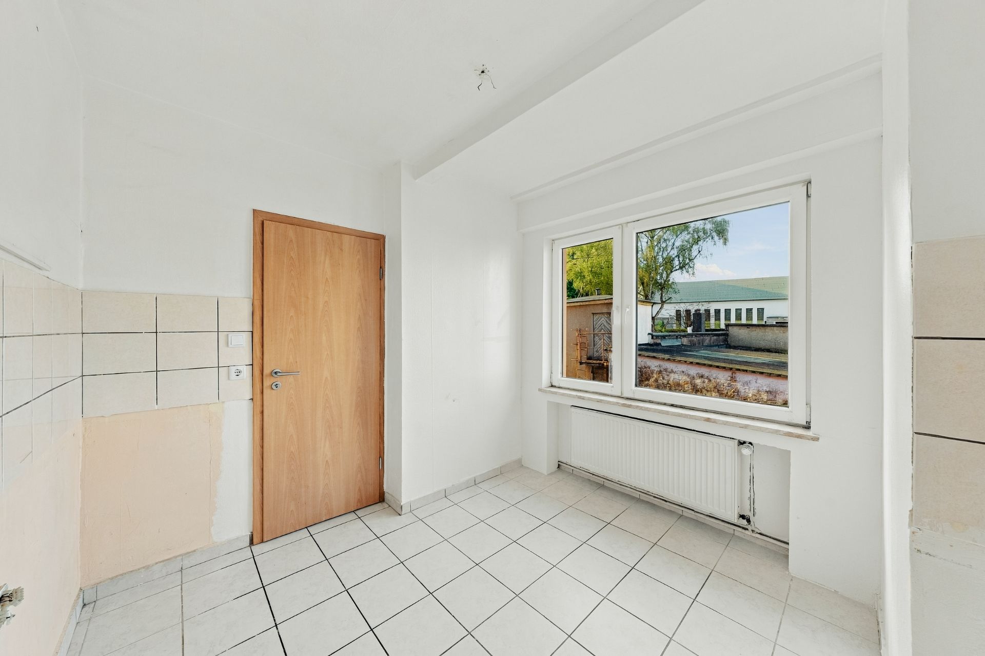 Heller, leerer Raum mit Fliesenboden, Holztür und großem Fenster mit Blick nach draußen.