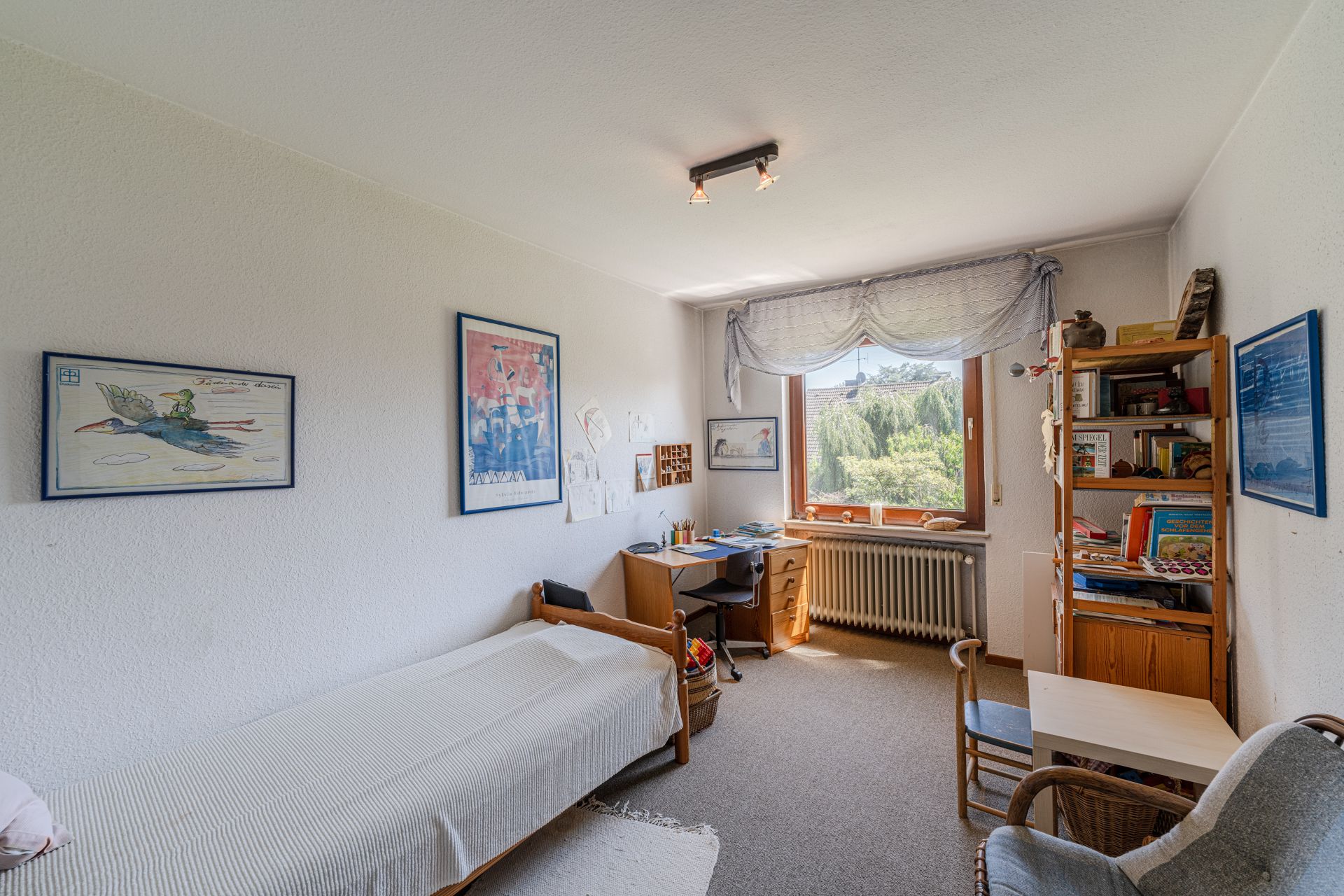 Helles Kinderzimmer mit Bett, Schreibtisch, Regal, Fenster und Wandbildern.