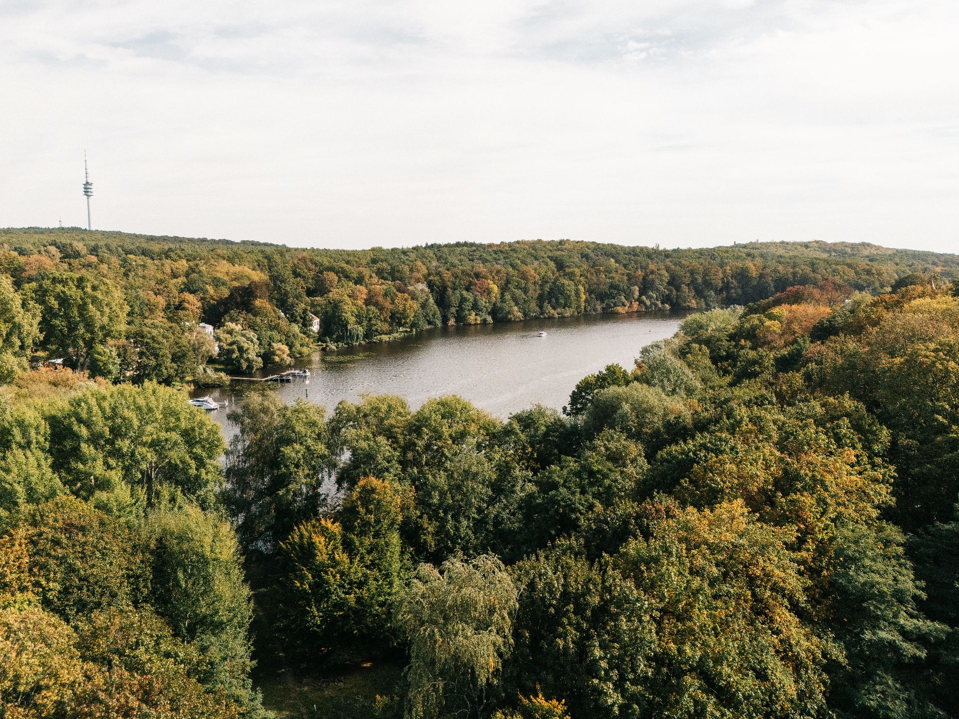 Blick auf einen bewaldeten See und umliegende Bäume unter bewölktem Himmel.