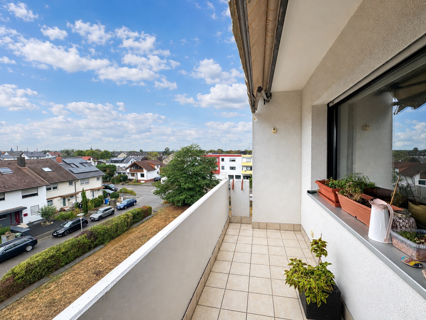 Balkon mit Pflanzkübeln, Blick auf Wohnhäuser und Straße unter blauem Himmel.