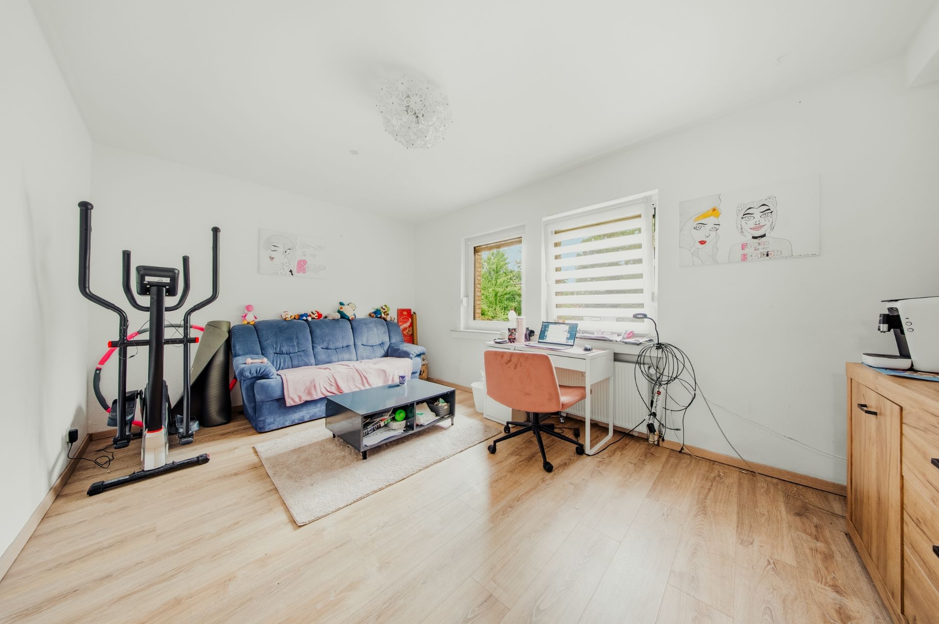 Helles Zimmer mit Sofa, Schreibtisch, Ergometer und Spielzeug auf Holzfußboden.