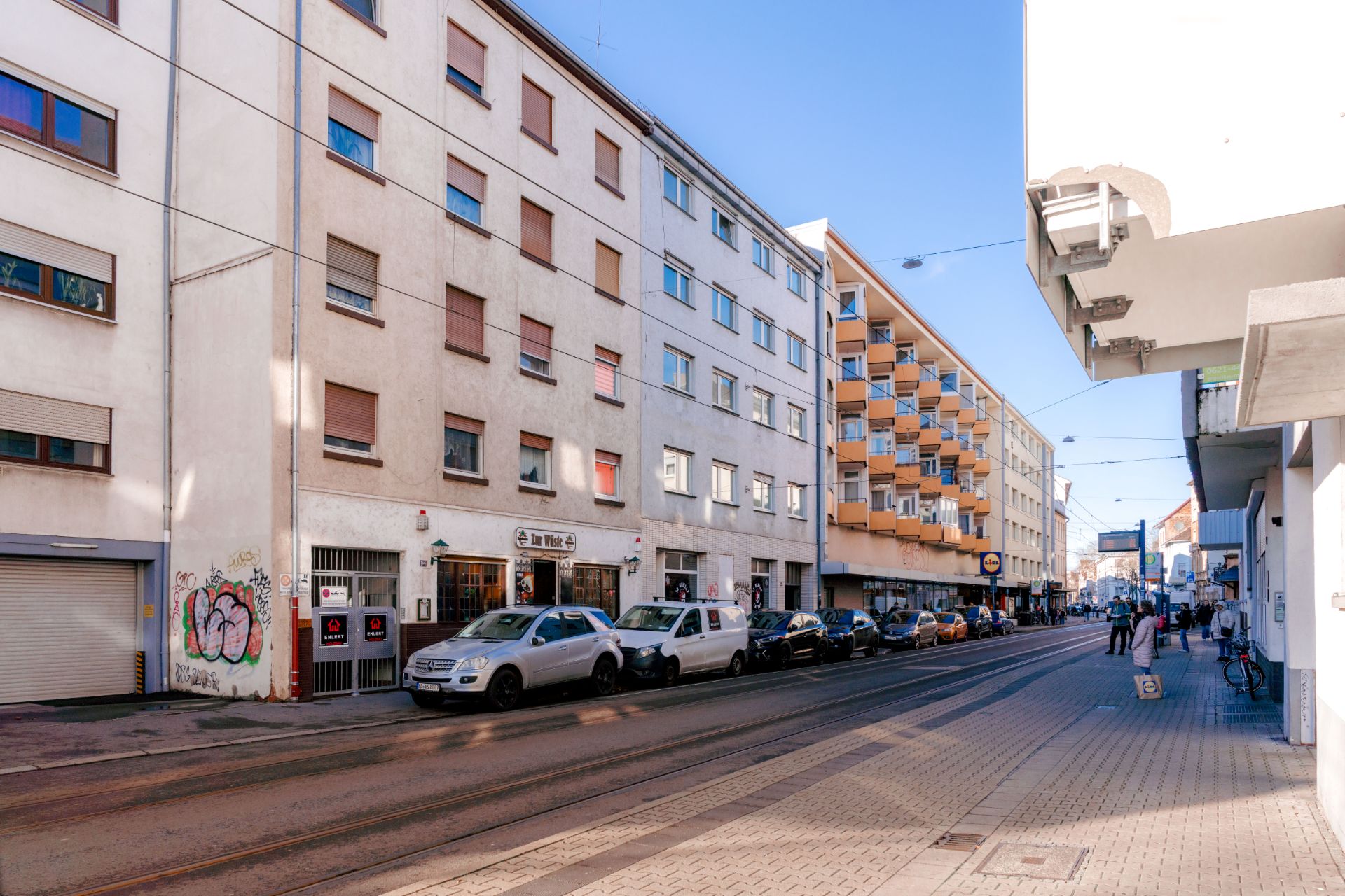 Mehrstöckiges Wohnhaus an einer Straße mit geparkten Autos und einigen Passanten auf dem Gehweg.