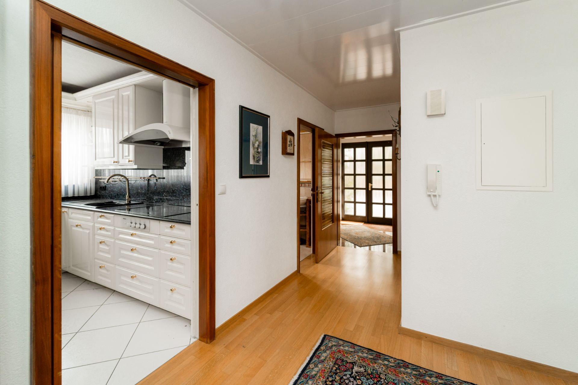 Heller Flur mit Holzboden, Zugang zur weißen Küche und Glastür zum Wohnzimmer.