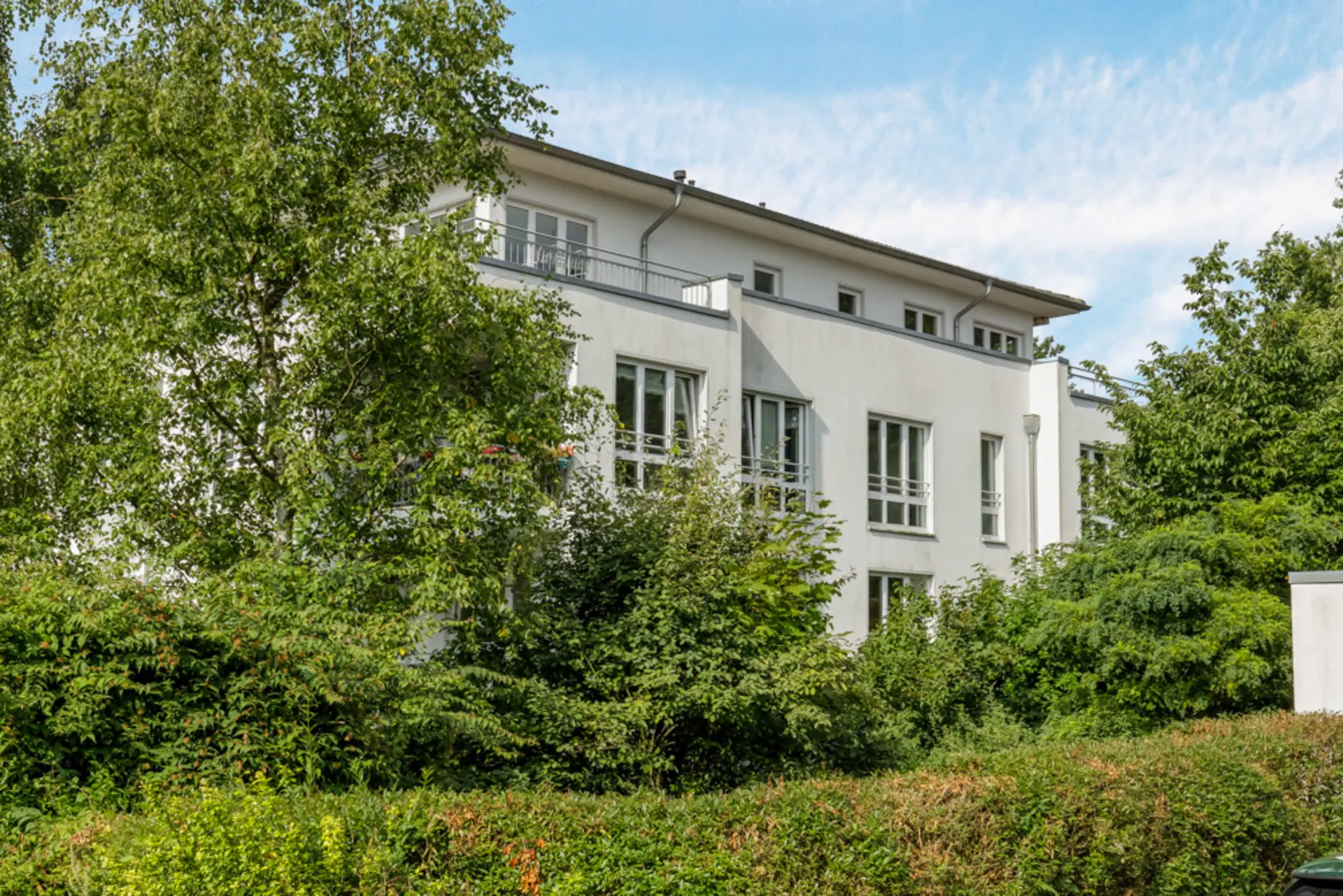 Weißes Wohngebäude mit großen Fenstern, umgeben von Bäumen und Büschen.