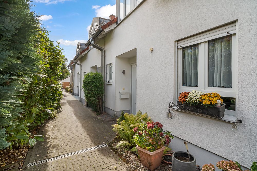 Gepflasterter Weg neben weißem Haus mit Blumen im Fensterkasten und Blumentöpfen am Eingang.