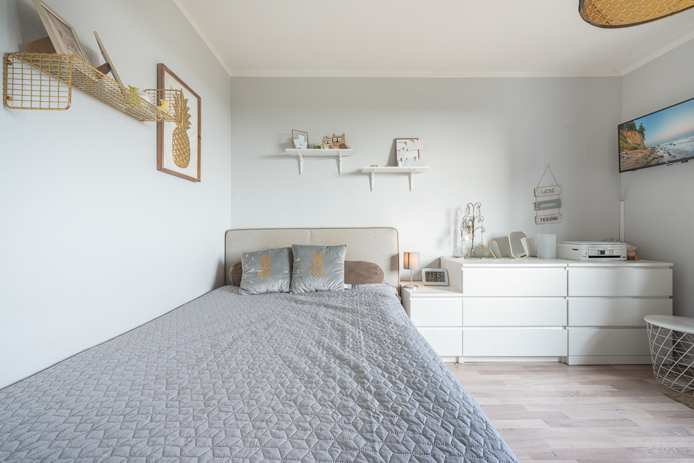 Helles Schlafzimmer mit grauer Bettdecke, weißen Möbeln und Dekoration an den Wänden.