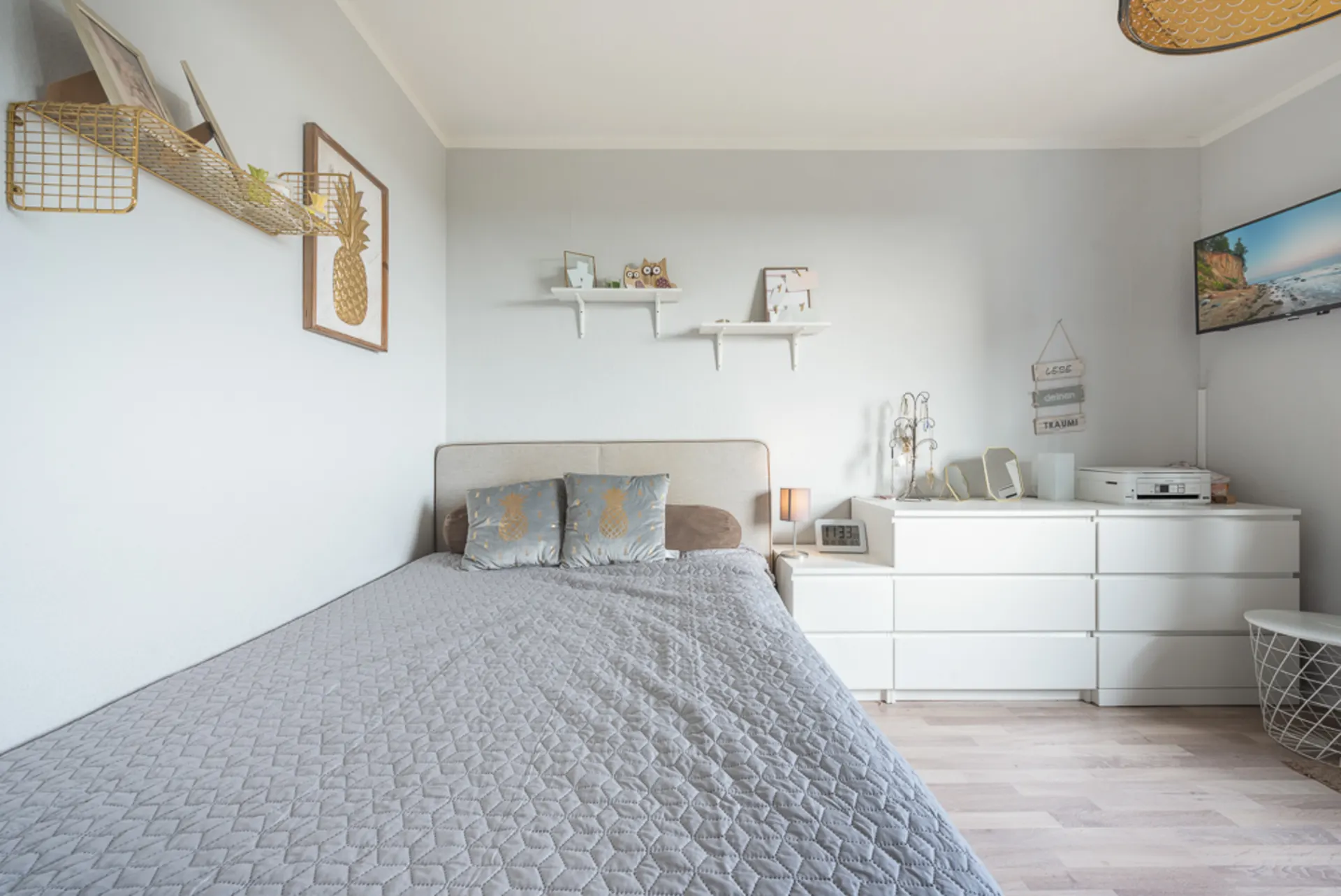Helles Schlafzimmer mit grauer Bettdecke, weißen Möbeln und Dekoration an den Wänden.