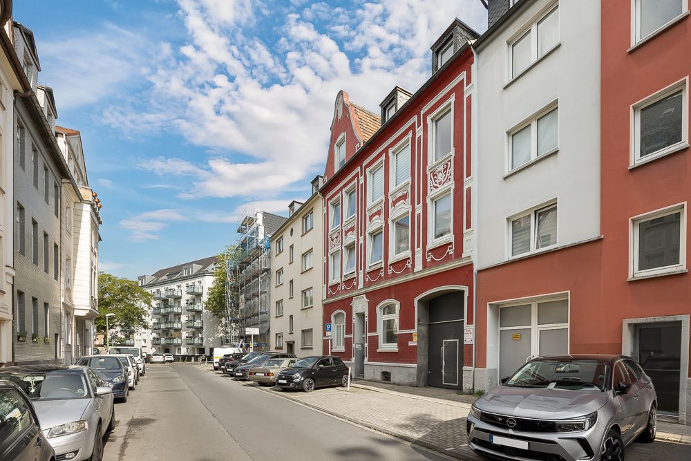 Wohnstraße mit parkenden Autos und mehrstöckigen Wohnhäusern, davon eines rot mit weißen Verzierungen.