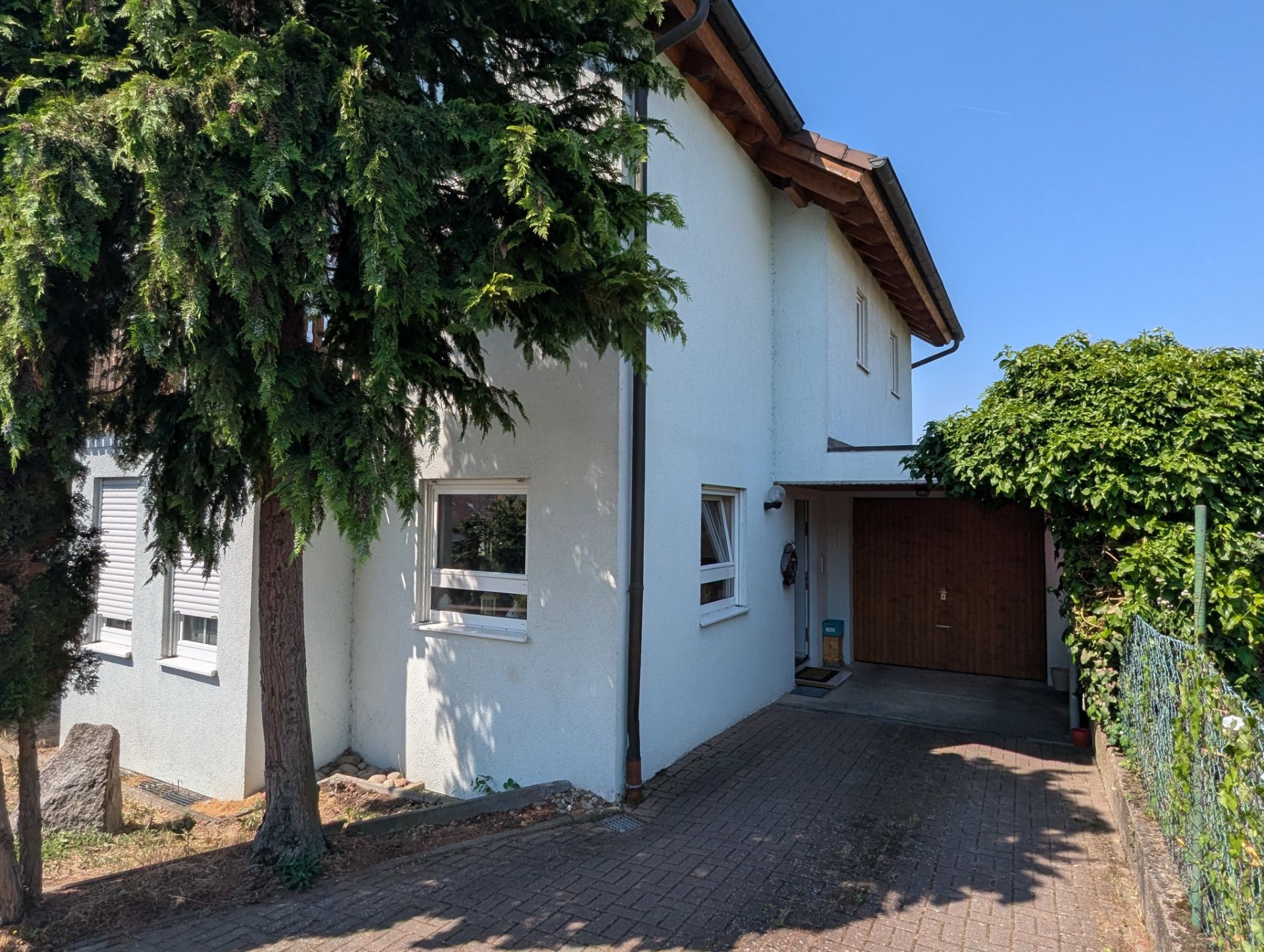 Weißes Wohnhaus mit Garage, Einfahrt und Bäumen im Vordergrund bei sonnigem Wetter.