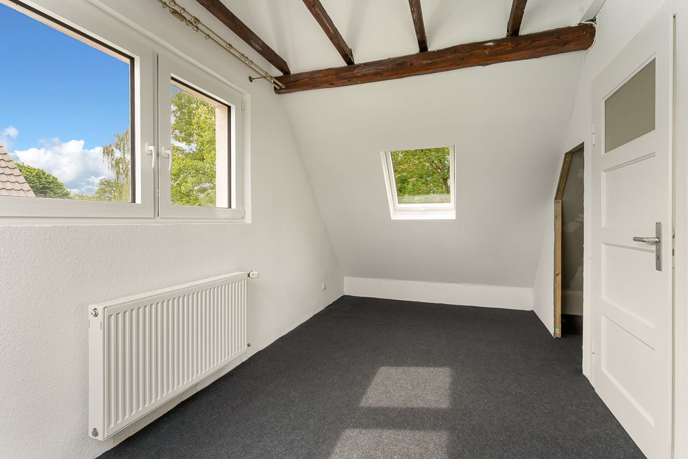 Helles Zimmer mit sichtbaren Holzbalken, zwei Fenstern, Tür und grauem Teppichboden.