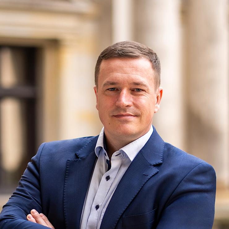 Ein Mann im blauen Anzug steht draußen mit verschränkten Armen.