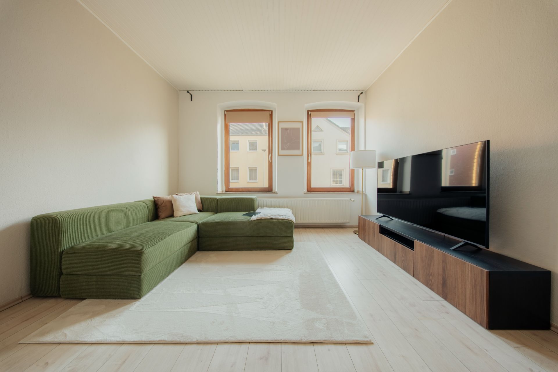 Wohnzimmer mit grünem Ecksofa, Teppich, Stehlampe und großem Fernseher vor zwei Fenstern.