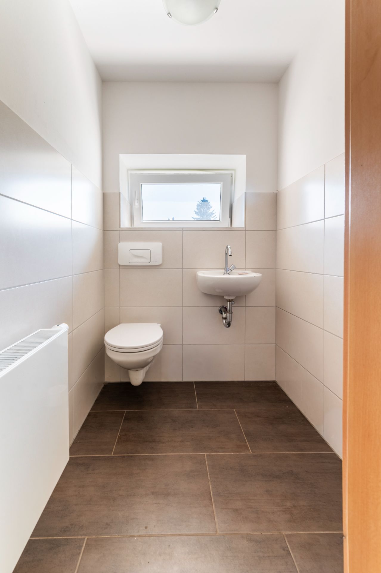 Heller, schmaler WC-Raum mit wandhängender Toilette, kleinem Waschbecken und Fenster.