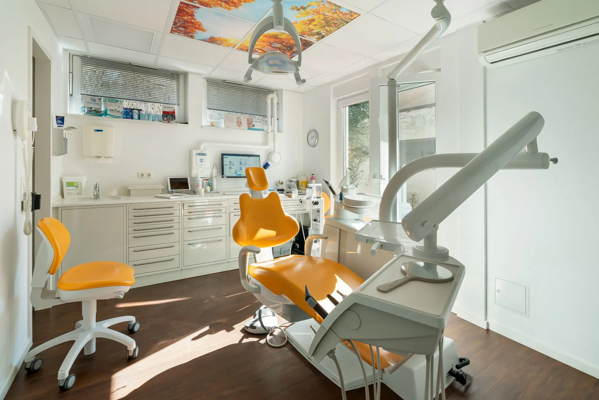 Heller Behandlungsraum mit orangefarbenem Zahnarztstuhl, Geräten und weißen Schränken.