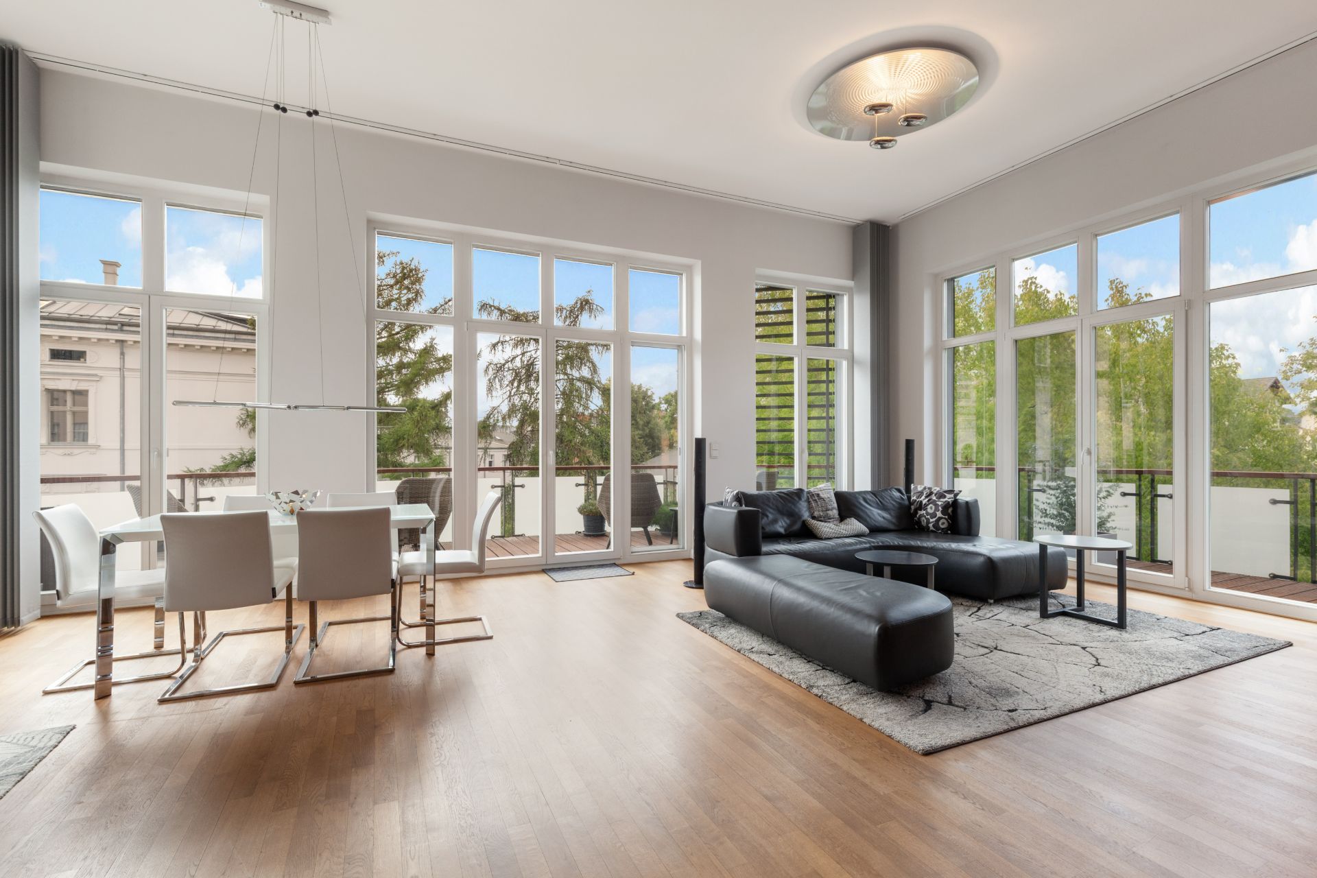 Helles Wohnzimmer mit großer Fensterfront, schwarzem Sofa, Esstisch und Holzboden.