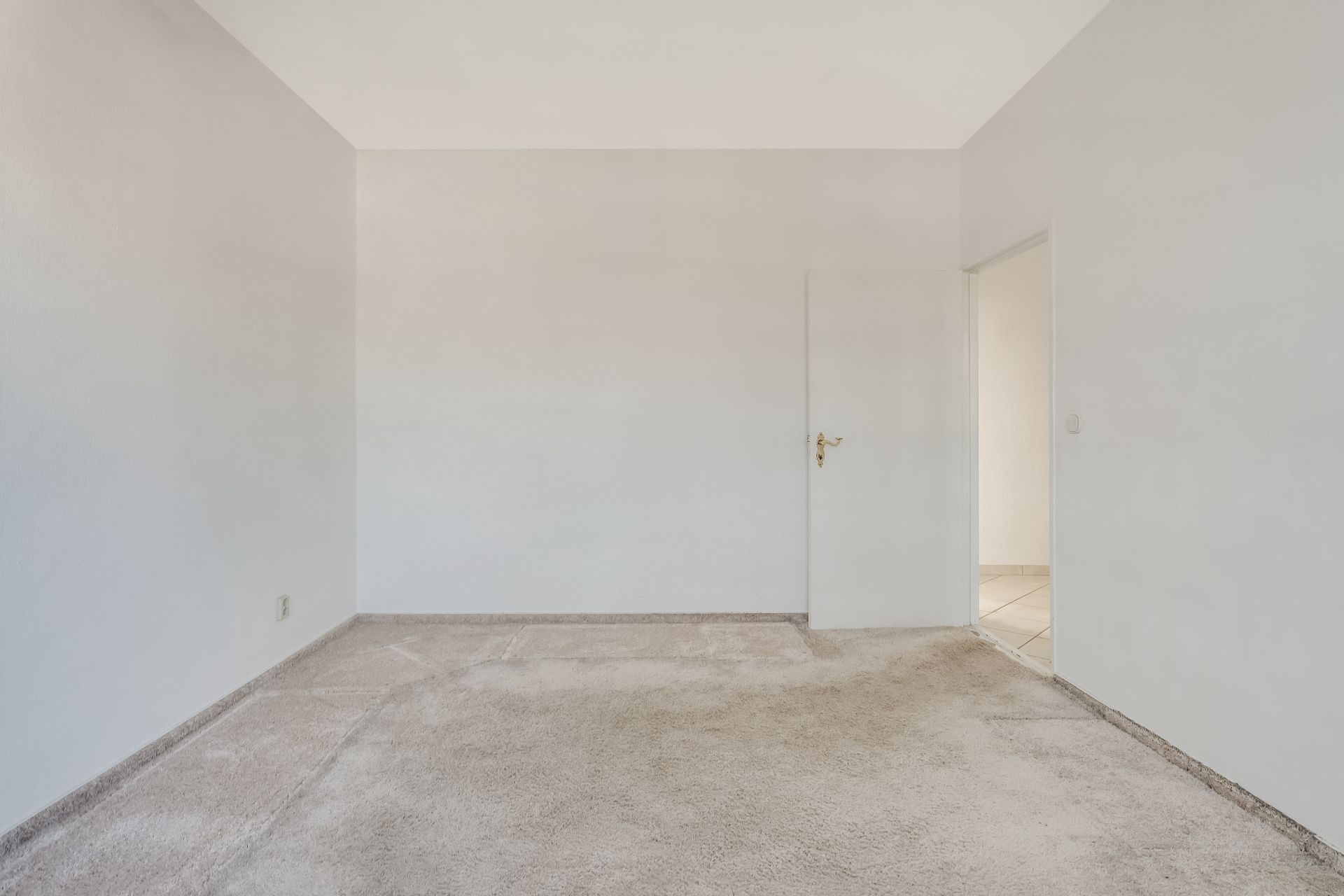 Leeres Zimmer mit weißem Teppichboden, weißen Wänden und geöffneter Tür.