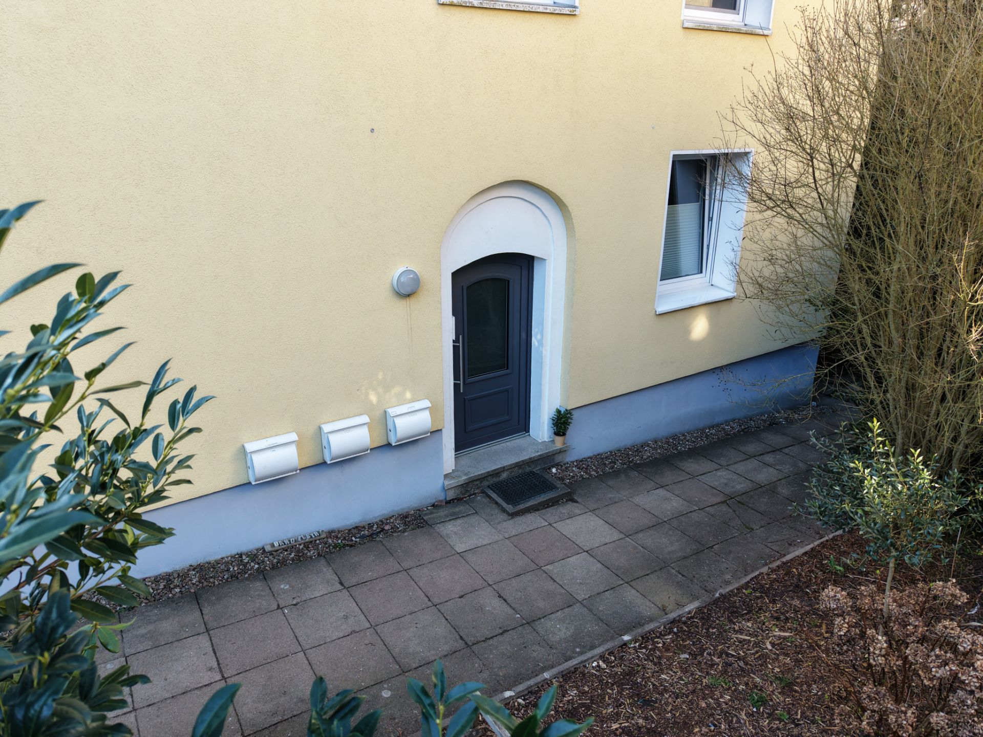 Haus­eingang mit grauer Tür, gelber Fassade und Fenster, Weg und Pflanzen im Vordergrund.
