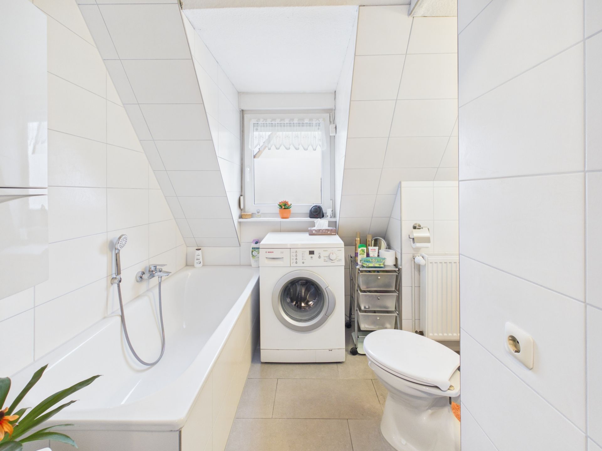 Helles Badezimmer mit Badewanne, Waschmaschine, WC und Fenster unter einer Dachschräge.