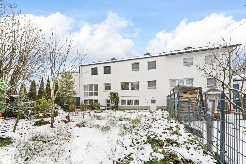 Weiße Wohnhäuser mit Garten im Winter, teils schneebedeckt, Bäume und Zaun im Vordergrund.