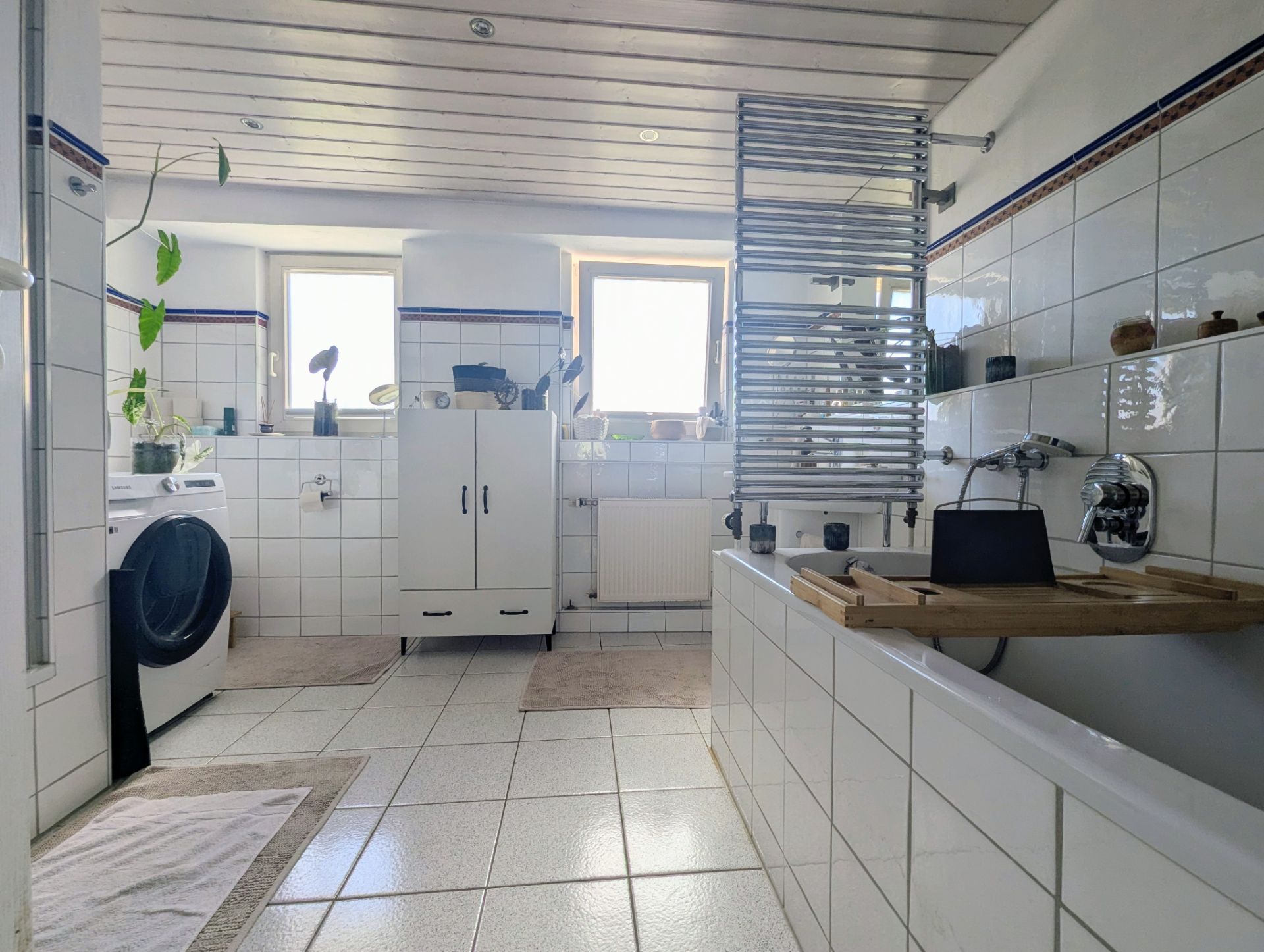 Modernes Badezimmer mit Badewanne, Waschmaschine, zwei Fenstern und weißen Fliesen.