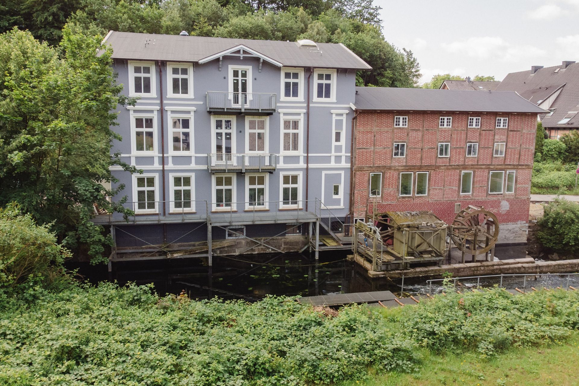 Zwei Wohngebäude am Wasser mit Wasserrad, eines blau-weiß, das andere aus rotem Backstein.
