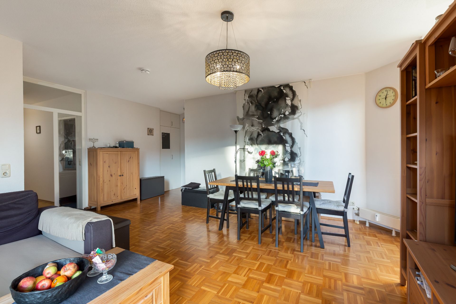 Helles Wohnzimmer mit Esstisch, sechs Stühlen, Sofa, Schrank und Wanduhr.