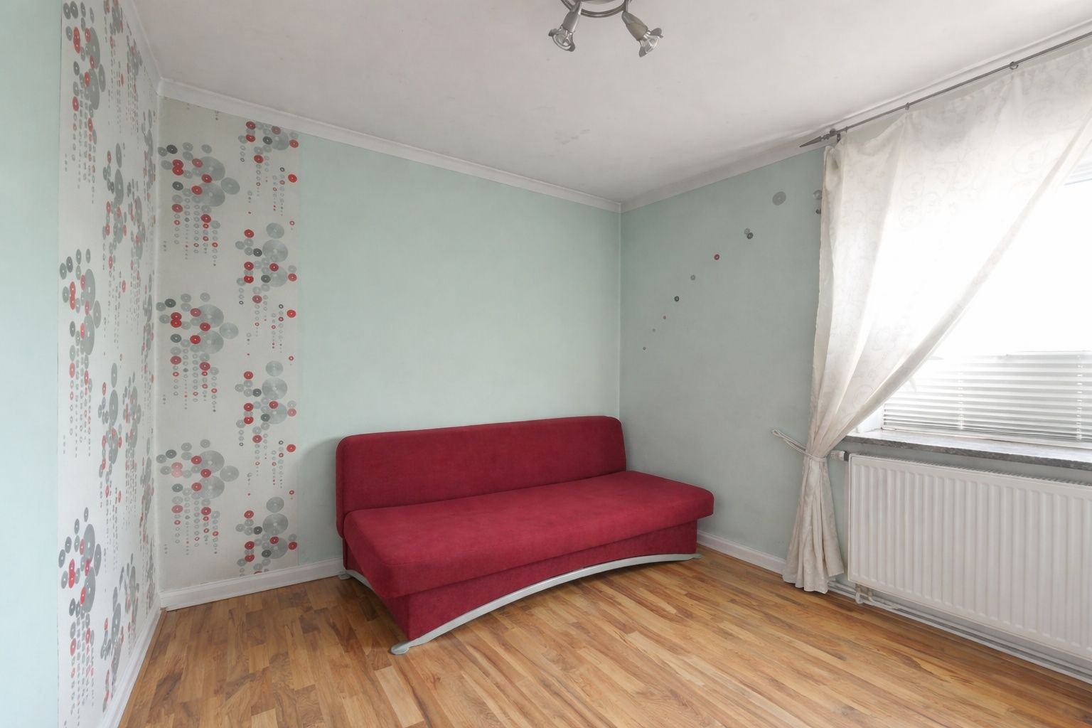 Kleines Zimmer mit rotem Sofa, Holzboden, gemusterten Wänden und großem Fenster mit Vorhang.