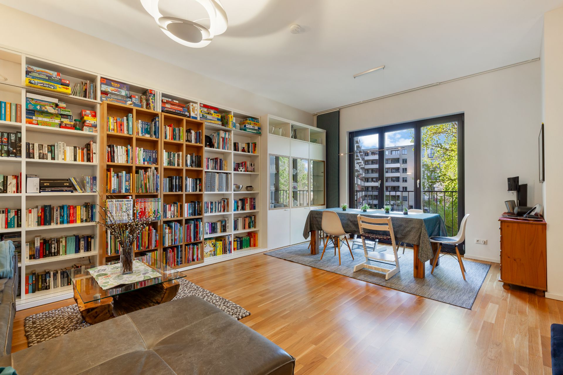 Wohnzimmer mit Bücherregal, Esstisch, großen Fenstern und Holzboden.