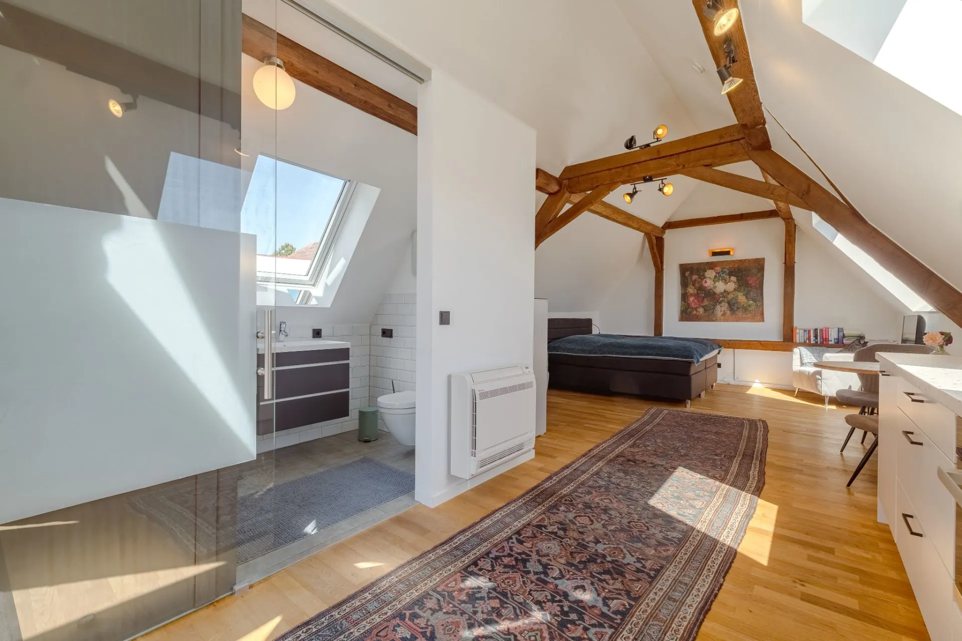 Helles Schlafzimmer mit sichtbaren Holzbalken, Teppich, Bett, Schreibtisch und offenem Badezimmer.