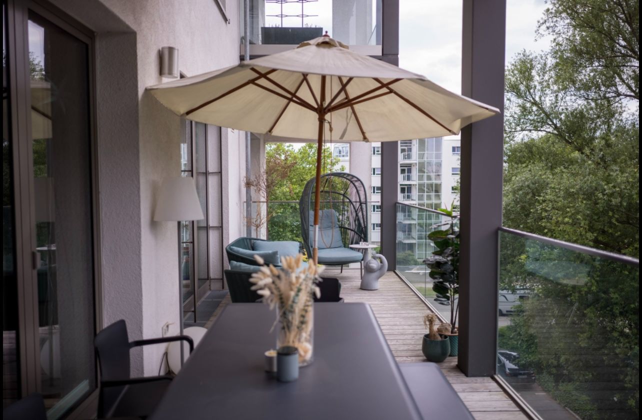 Balkon mit Tisch, Stühlen, großem Sonnenschirm und Lounge-Bereich, Blick auf Bäume und andere Gebäude.