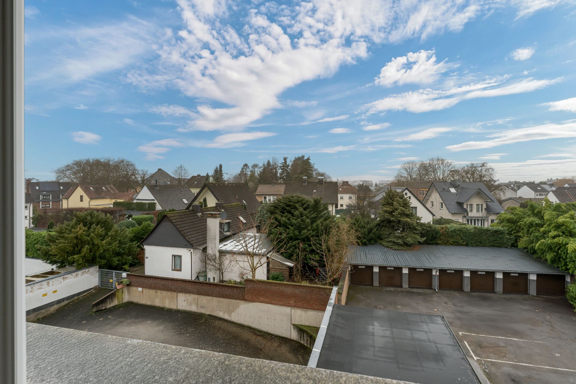 Blick aus dem Fenster auf Wohnhäuser, Garagen und einen Hof bei blauem Himmel mit Wolken.