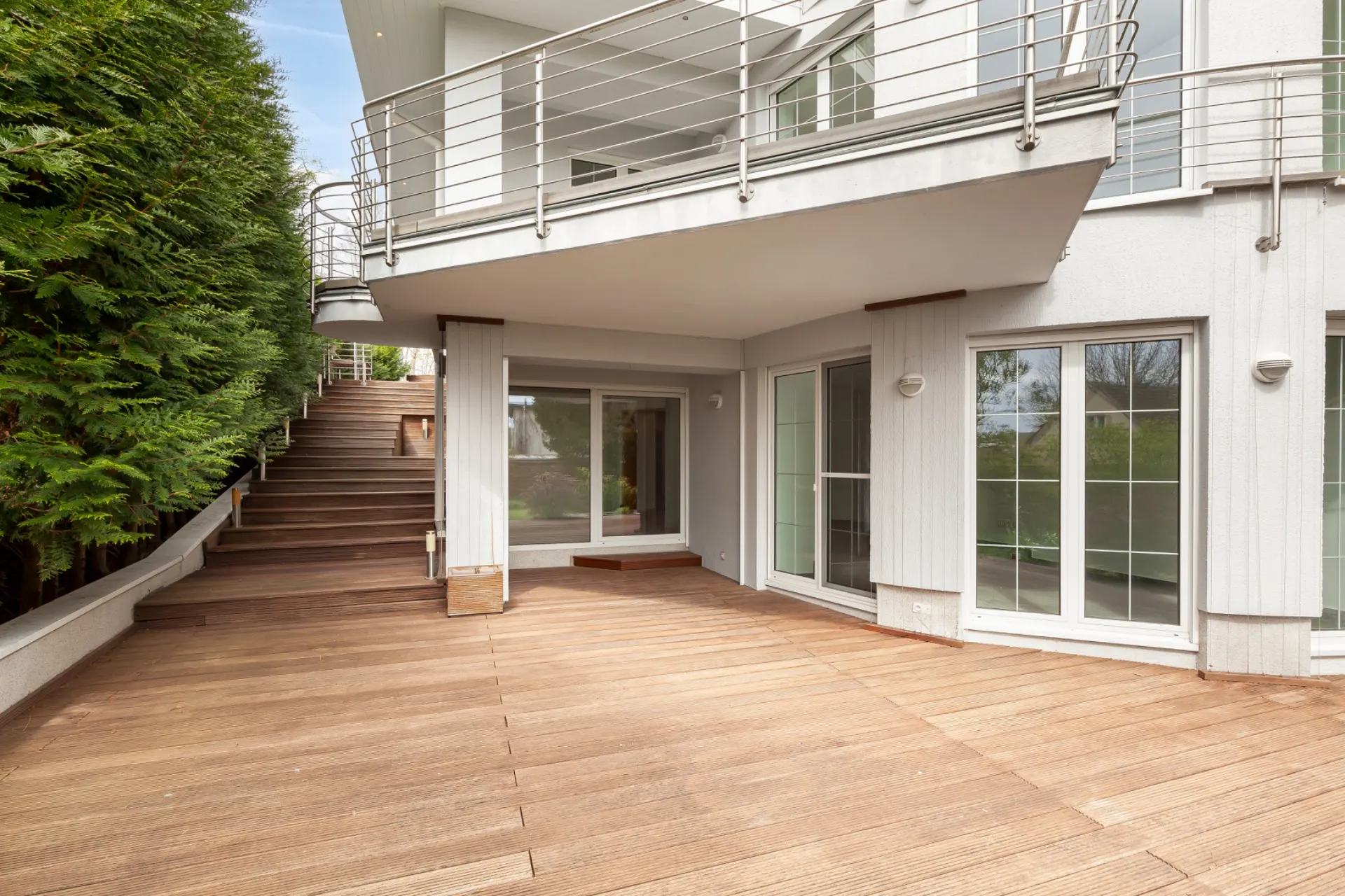 Große Terrasse mit Holzboden, seitlicher Treppe und weißen Fenstern an einem Wohnhaus.