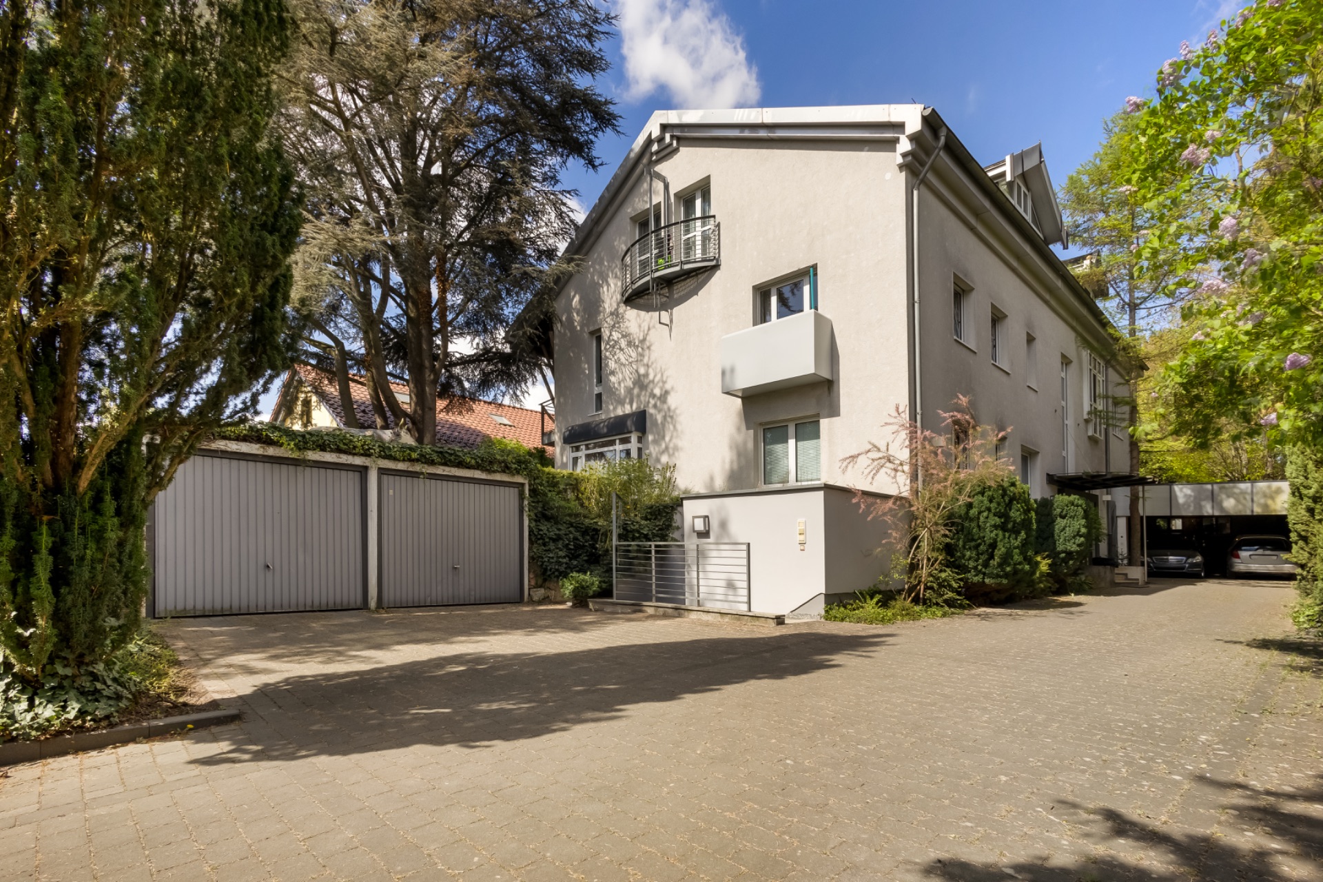 Mehrfamilienhaus mit Garagen, Bäumen und gepflasterter Einfahrt bei Tageslicht.