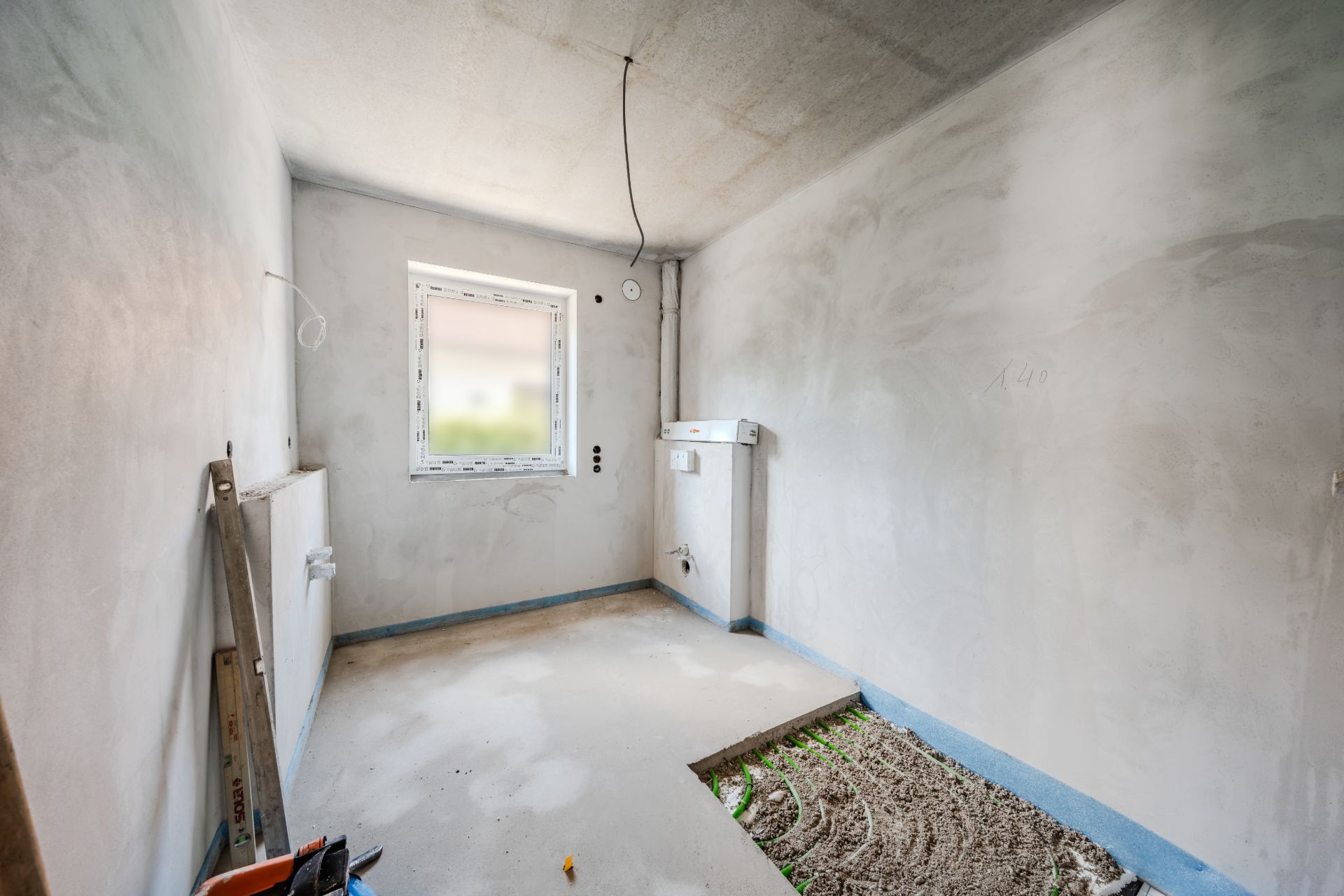 Leerer Raum mit verputzten Wänden, einem Fenster und offenen Leitungen am Boden.