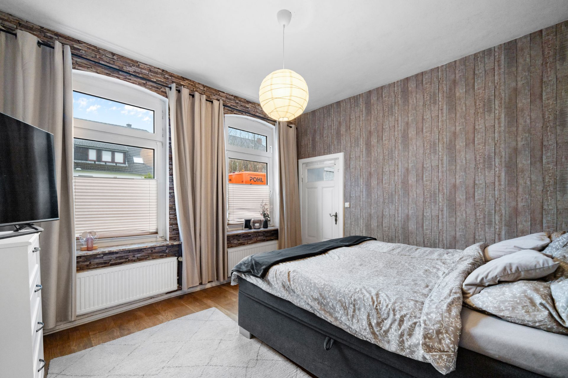 Helles Schlafzimmer mit Doppelbett, TV, zwei Fenstern und grauen Vorhängen.