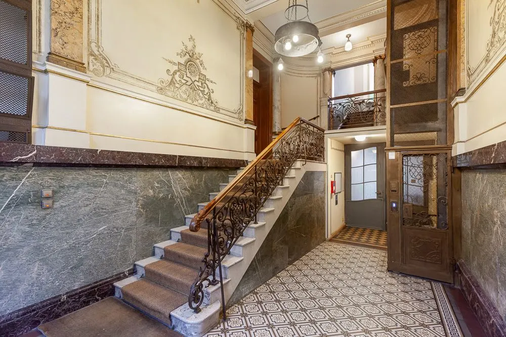 Eingangsbereich mit Treppe, schmiedeeisernem Geländer, Aufzug und gemustertem Fliesenboden.