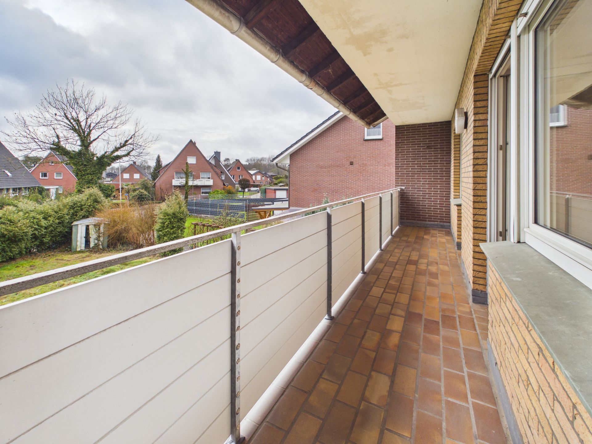 Langer Balkon mit braunen Fliesen an einem Wohnhaus, Blick auf Nachbarhäuser und Gärten.
