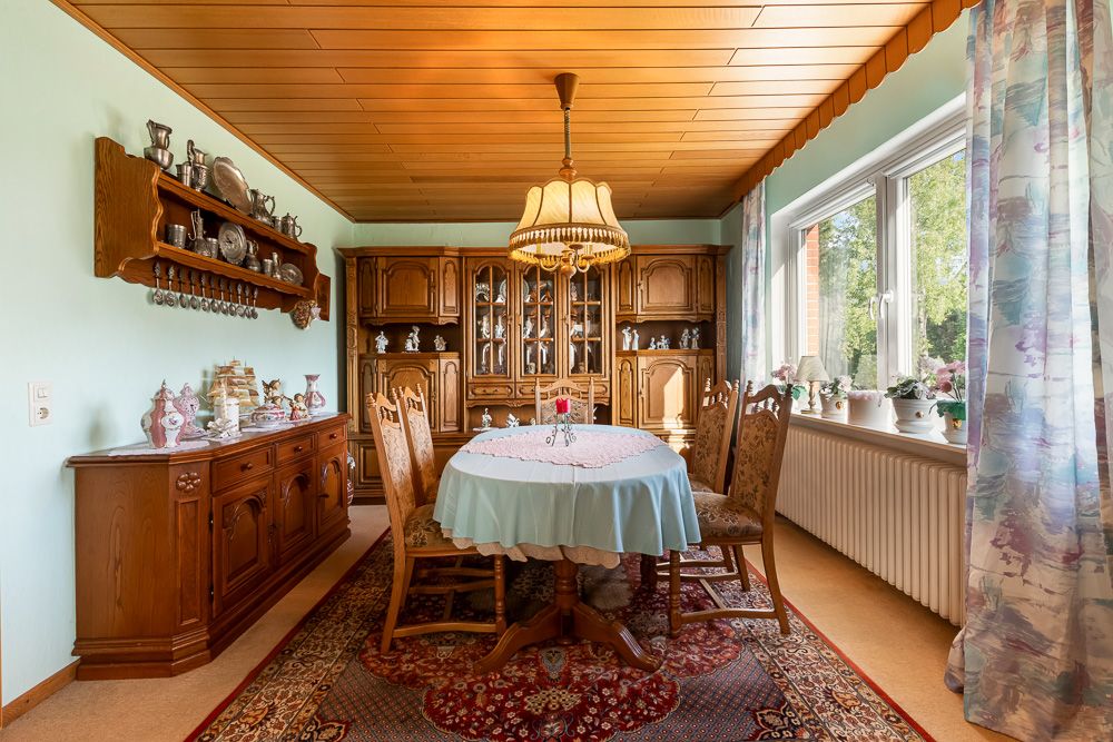 Gemütliches Esszimmer mit Holzmöbeln, rundem Tisch, Teppich und großem Fenster mit Gardinen.